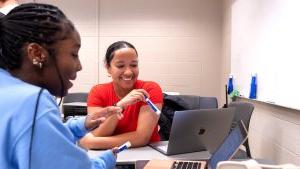 A student tutor sitting a desk with a laptop tutoring a student who also has her laptop out.