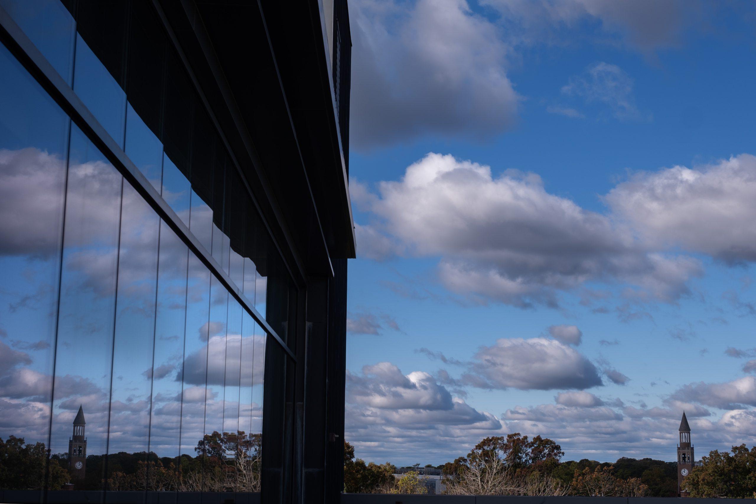 Long-distance shot of the Morehead-Patterson Bell Tower on the campus of U.N.C Chapel Hill on a sunny day. Clouds are seen in the background, as is a reflection of the scene, bouncing off the windows of a building.